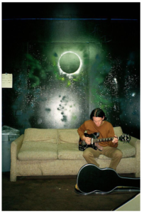 Elliott Smith playing guitar while sitting on a couch.